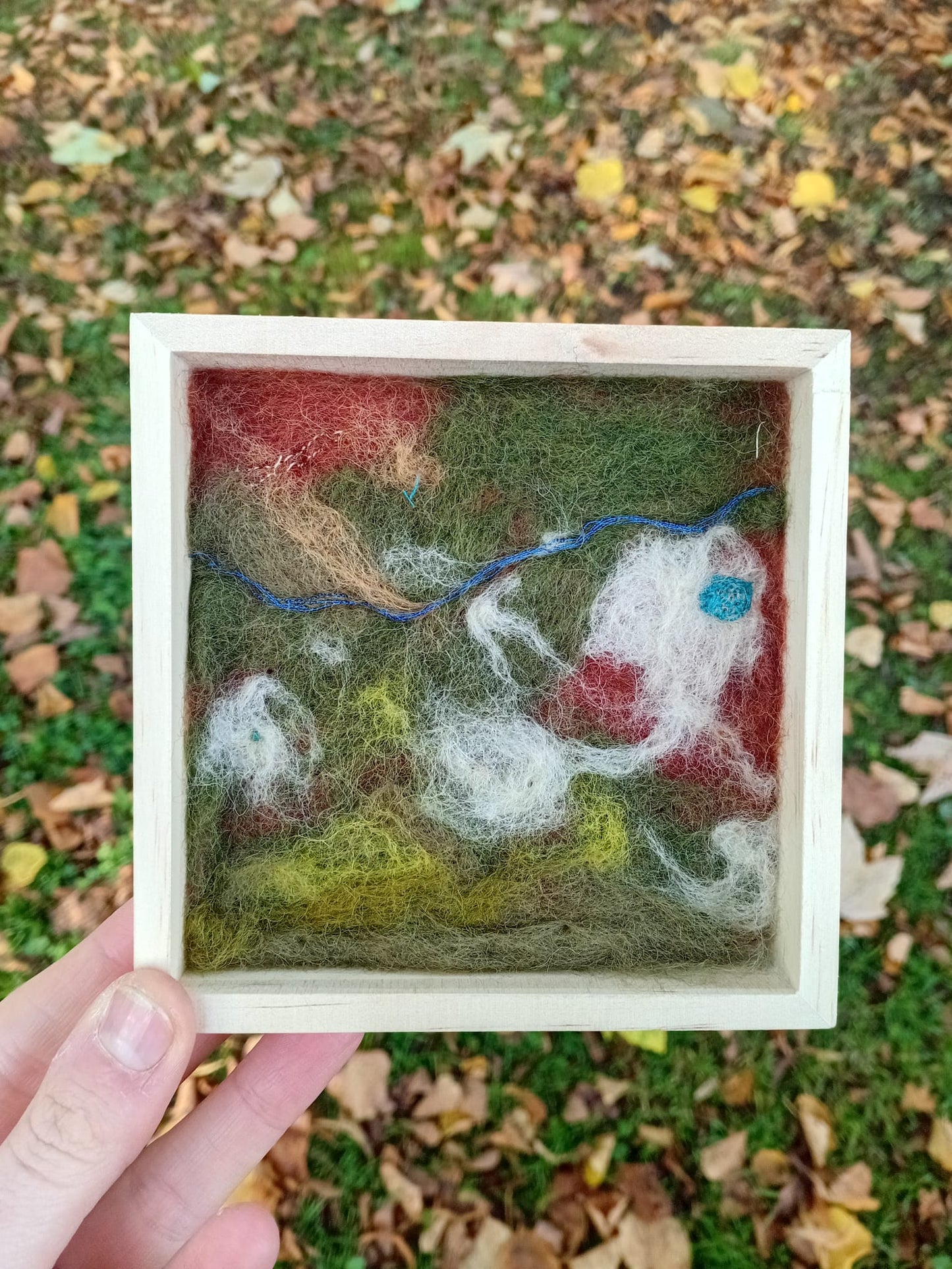 Felted artwork of a landscape with animals in a wooden frame, held outdoors.