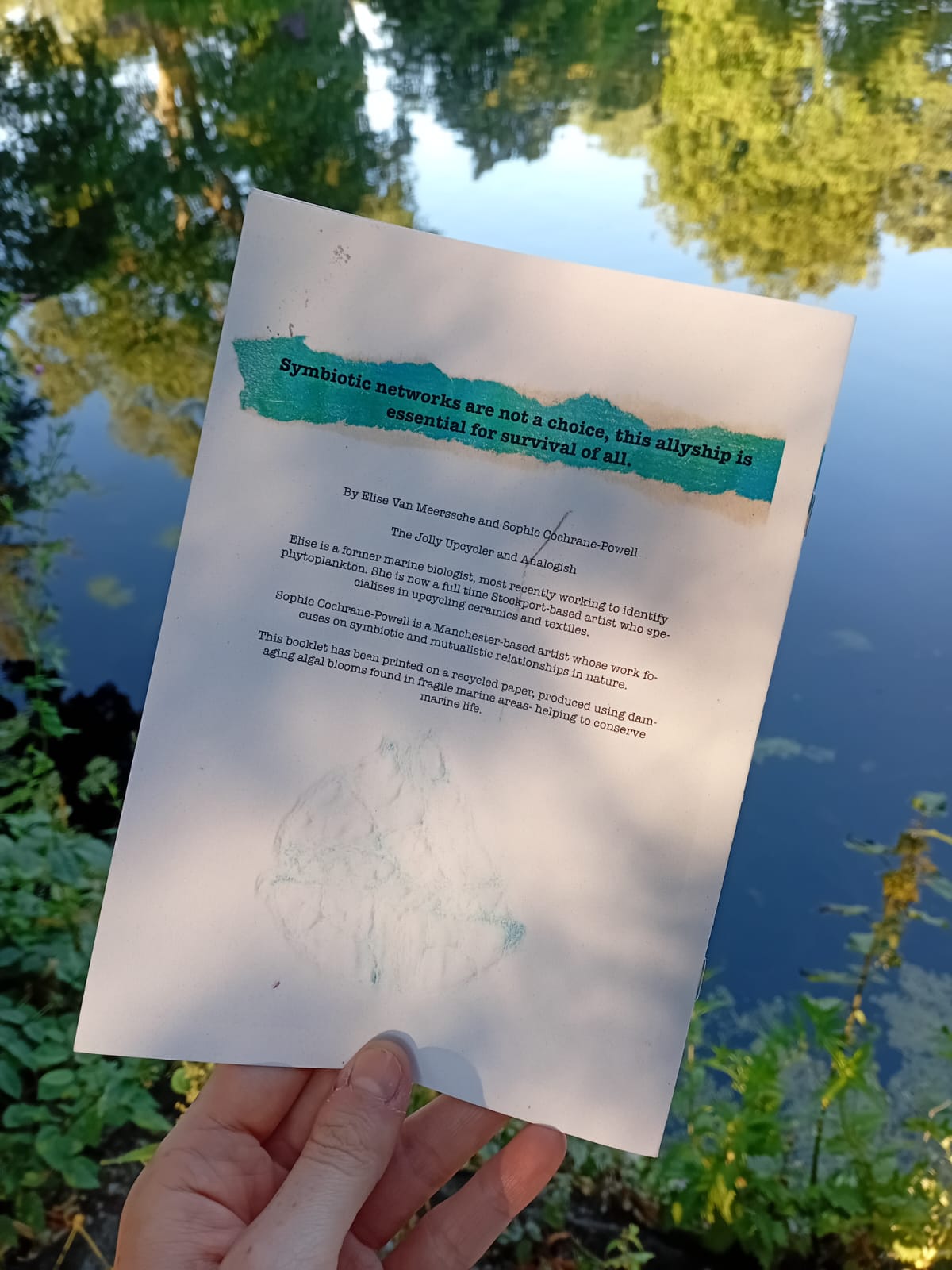 Hand holding a paper with text over a reflective water surface with trees.