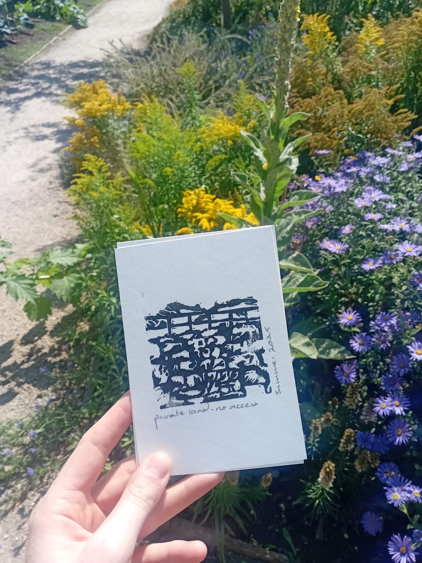 Hand holding a white card with black text against a background of flowers. Taken at Quarry Bank Mill National Trust Park
