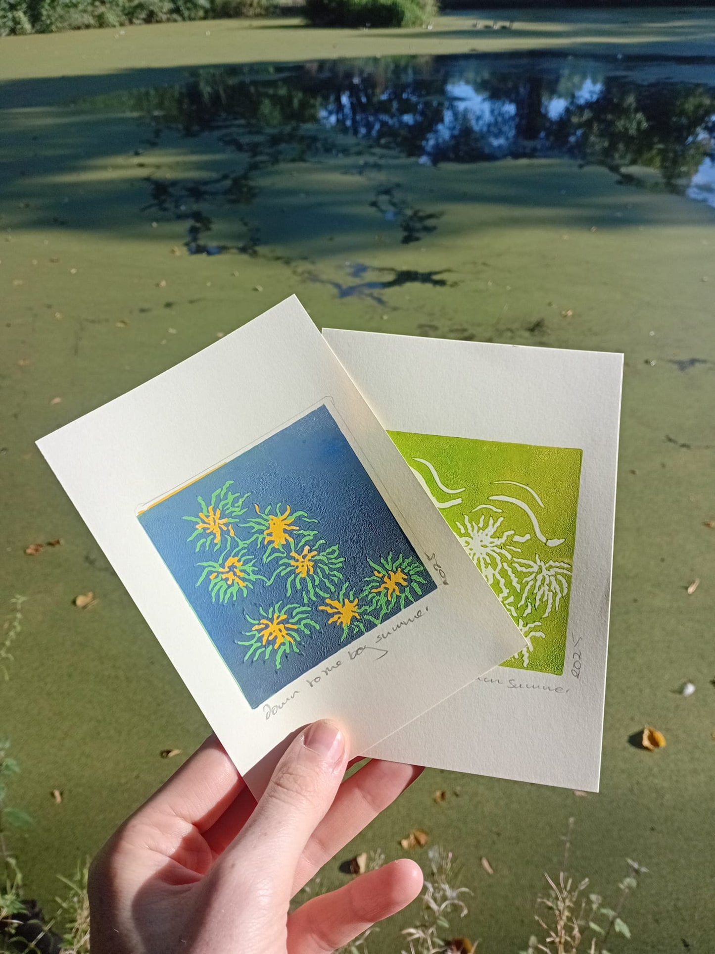 Two A6 lino prints held by a hand with a natural background of Alexandra Park in Whalley Range/ Moss Side, Manchester.