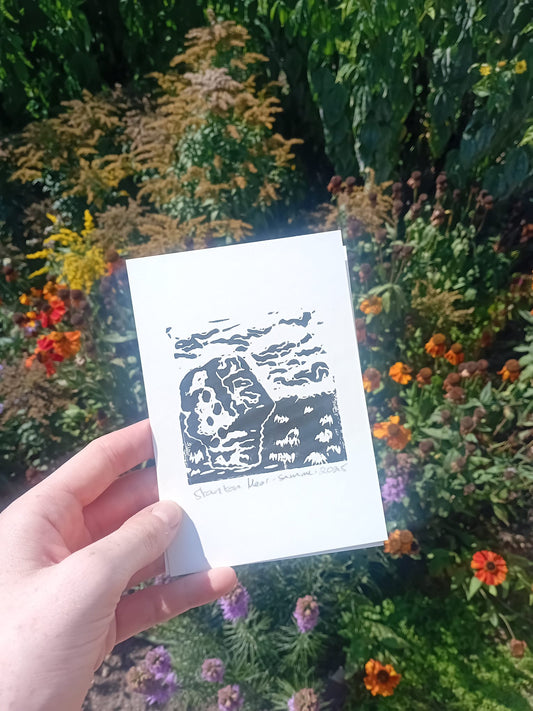Hand holding a small white card with black design against a floral background. The print is of the Andle Stone on Stanton Moor.