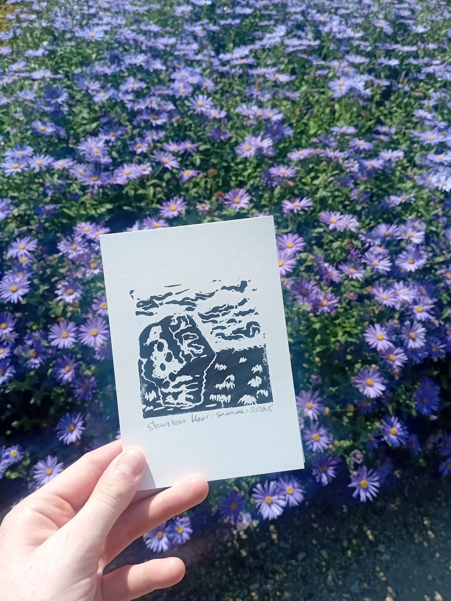 Hand holding a card with a black and white design against a background of purple flowers. Made by local Manchester-based artist during a trip to Matlock.