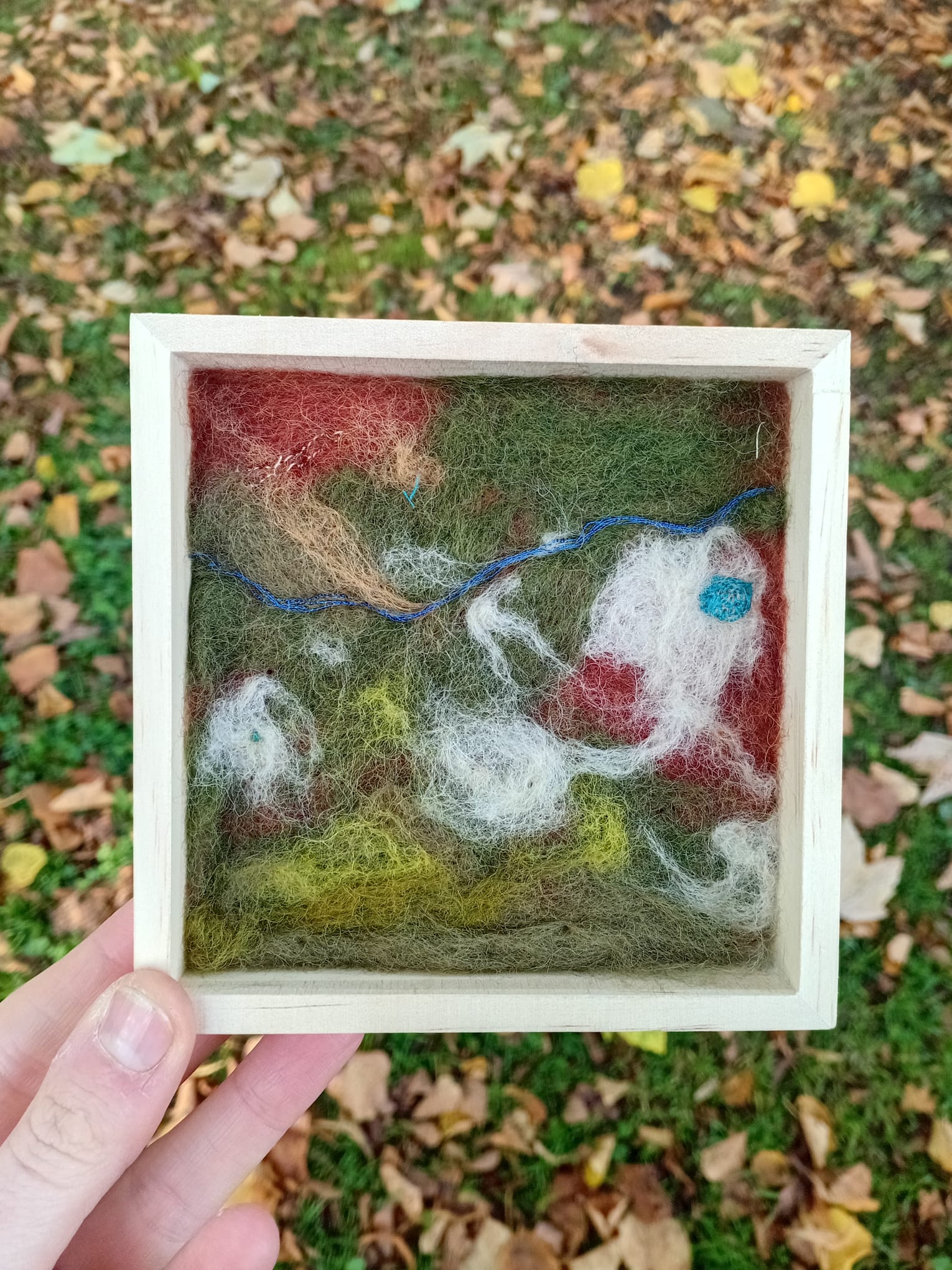 Felted artwork of a landscape with animals in a wooden frame, held outdoors.