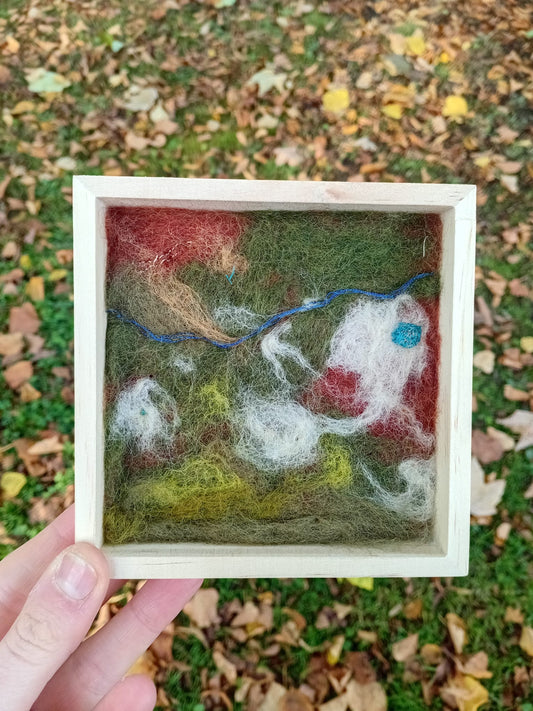 Felted artwork of a landscape with animals in a wooden frame, held outdoors.