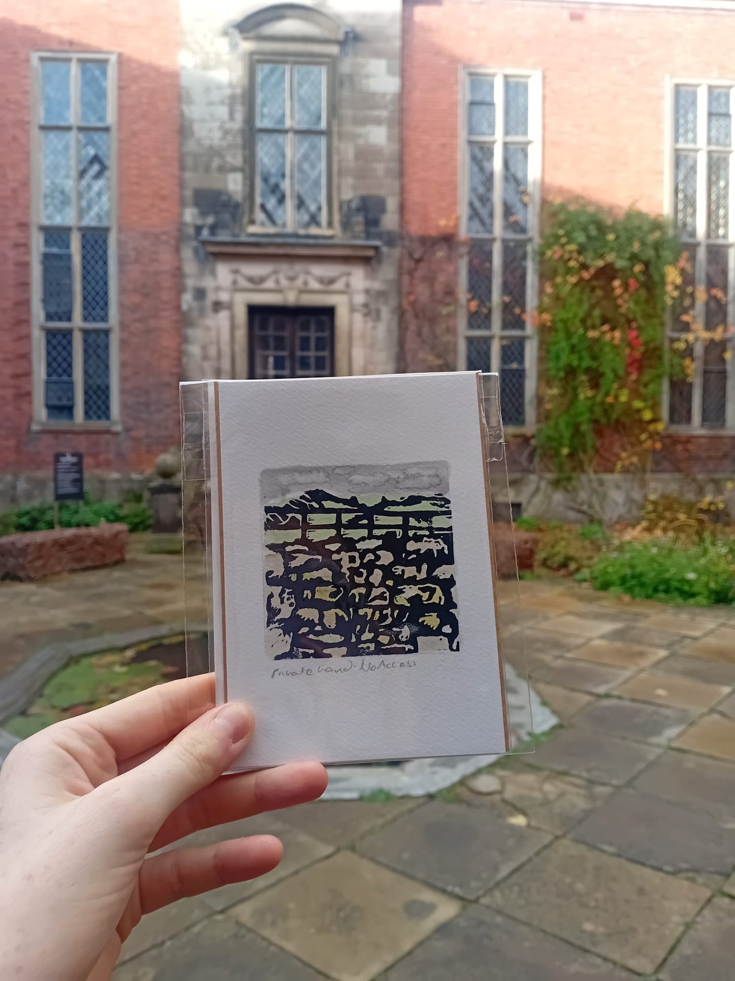 Hand holding a card with a patterned design in front of Dunham Massey Hall with large windows.