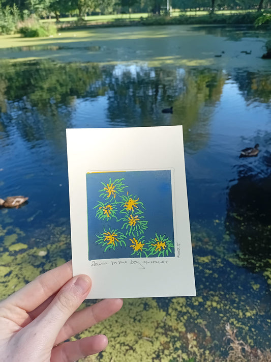 Hand holding a card with a floral design in front of a pond with ducks. The lino print has been made in Manchester and this park is Alexandra Park.