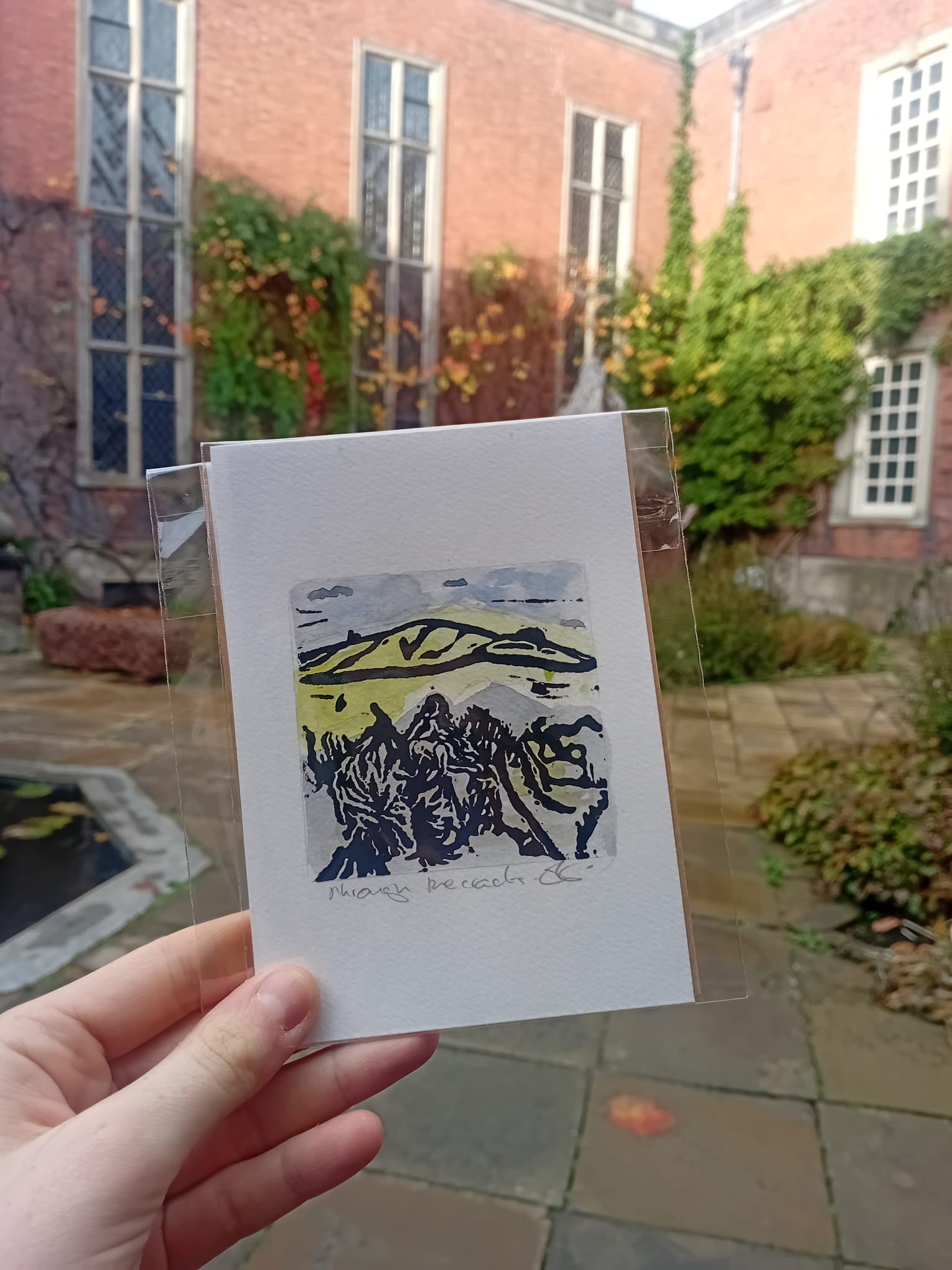 Hand holding a small print of a mountain landscape with a garden and building in the background.